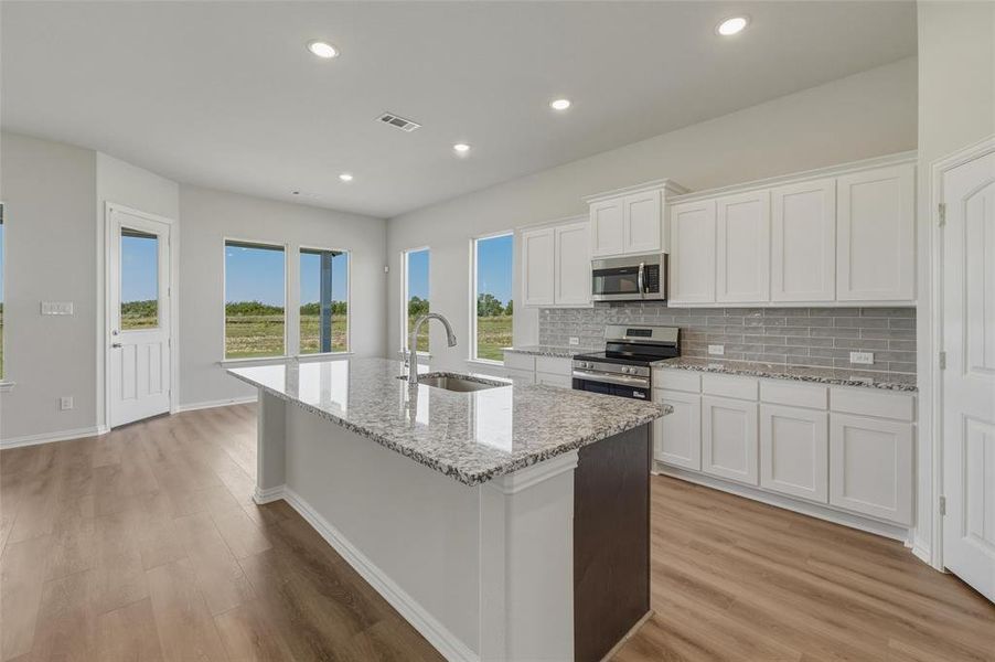Kitchen featuring a large central island with a granite countertop and undermount sink, stainless steel appliances, white cabinetry, and wood-finish flooring Kitchen featuring a large central island with a granite countertop and undermount sink, stainless steel appliances, white cabinetry, and wood-finish flooring