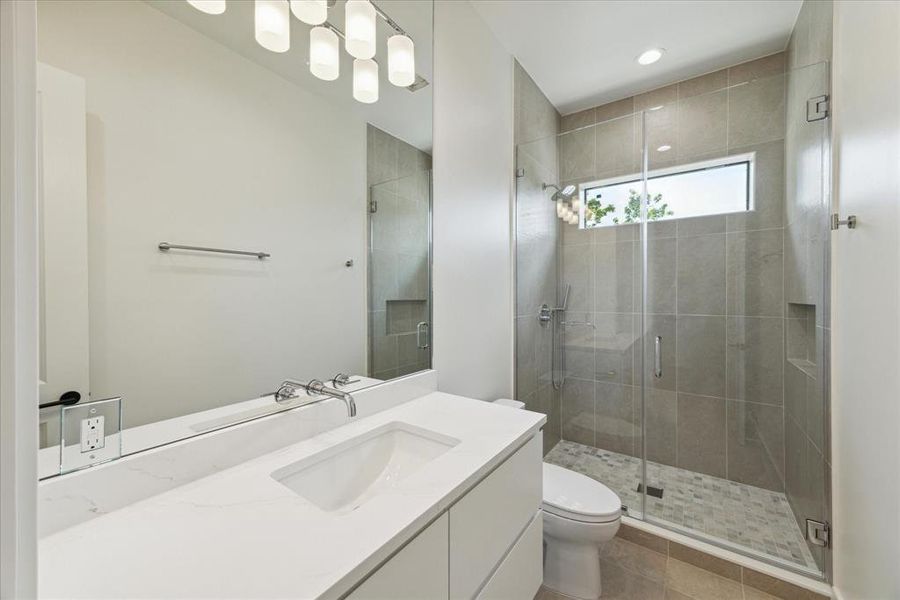 The ensuite bathroom for this bedroom features a walk-in shower with designer grey tile, a frameless glass door, and chrome plumbing fixtures. It includes quartz countertops, designer porcelain floor tile, and a custom vanity with chrome cabinet hardware. Additional highlights are a wall-mounted designer chrome faucet, an under-mount sink, and a counter-to-ceiling mirror.