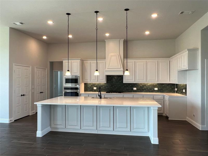 Kitchen with white cabinets, decorative light fixtures, a large island with sink, dark wood-type flooring, and light stone counters