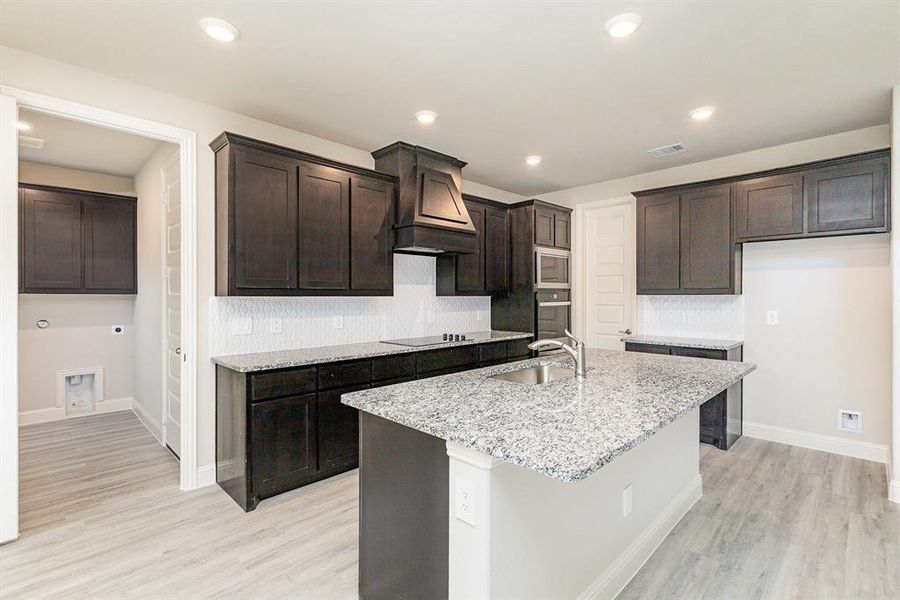 Kitchen featuring dark brown cabinetry, sink, light stone counters, an island with sink, and light hardwood / wood-style floors