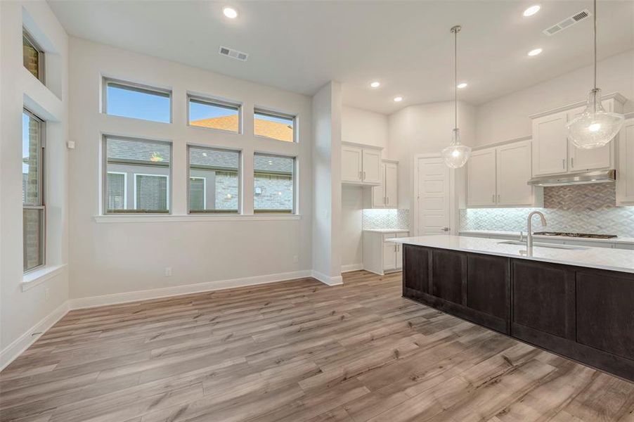 Two tone kitchen featuring backsplash, dual tone cabinetry, pendant lighting, light wood finished floors, and light stone counters
