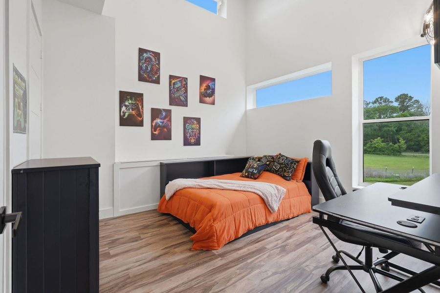 Bright, modern bedroom with high ceilings and large windows offering scenic views. Features stylish wall art, a comfortable bed with orange bedding, and a sleek desk setup, ideal for work or study.