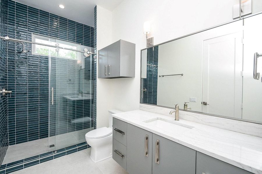 This modern bathroom features sleek gray cabinetry, a large mirror, and a stylish glass-enclosed shower with dark blue tiles. The space is bright and contemporary, offering a clean and functional design.