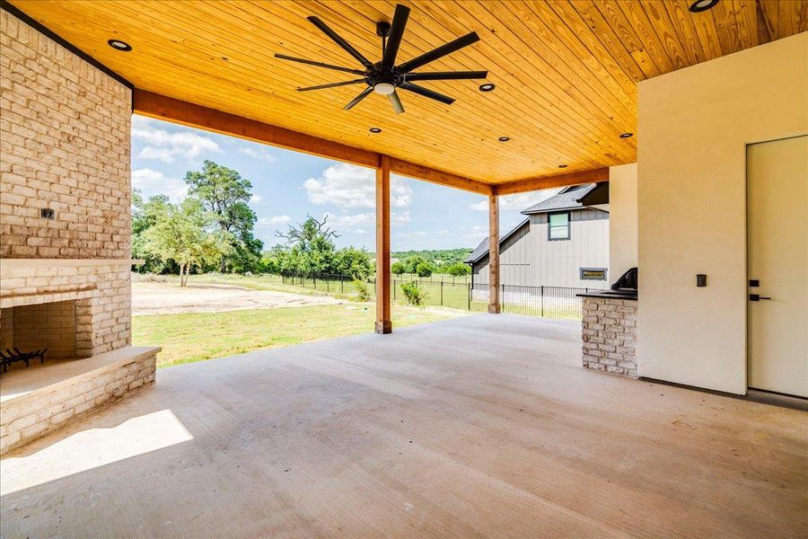 An inviting covered patio features a stone fireplace, outdoor kitchen, and a natural wood plank ceiling with recessed lighting.