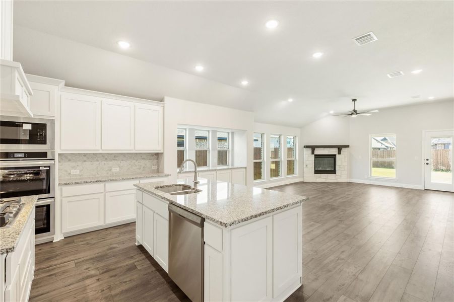 Open kitchen with white cabinets, granite island, and stainless appliances opening to living room with fireplace
