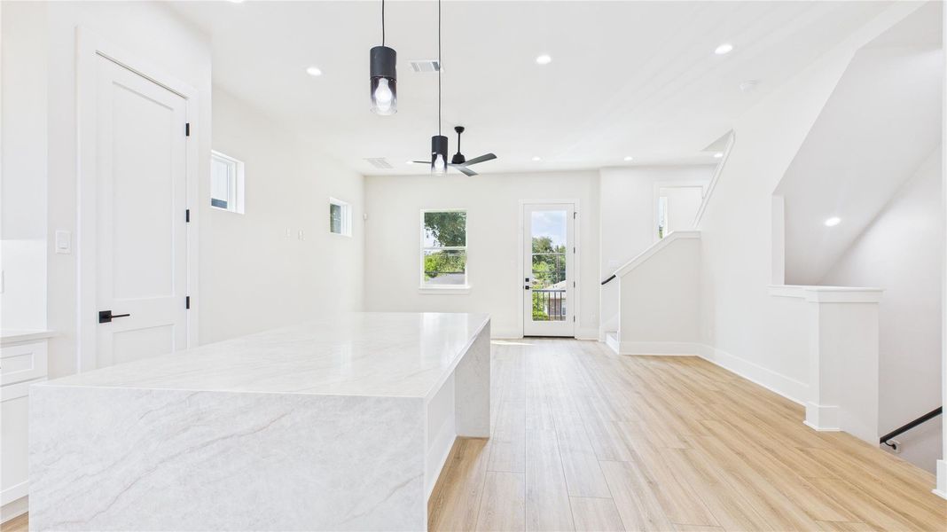 This bright, open-concept space features a modern design with light wood flooring, a large marble island, and ample natural light from multiple windows. The room includes recessed lighting and access to a small balcony, creating a welcoming and airy atmosphere.