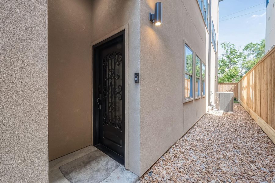 This is a side entry to a modern home featuring a secure black door with ornamental ironwork, a clean stucco exterior, and large windows for natural light. The area is neatly landscaped with pebbles and a wooden fence, providing privacy and a tidy appearance. This is a side entry to a modern home featuring a secure black door with ornamental ironwork, a clean stucco exterior, and large windows for natural light. The area is neatly landscaped with pebbles and a wooden fence, providing privacy and a tidy appearance.