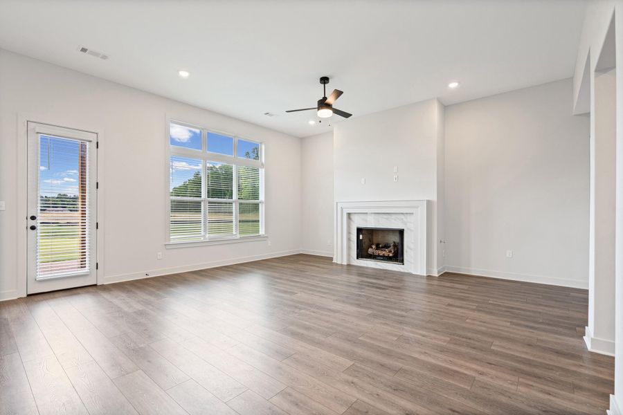 Spacious living area featuring wood-finish flooring, a white mantel fireplace with a stone surround, and abundant natural light from large windows Spacious living area featuring wood-finish flooring, a white mantel fireplace with a stone surround, and abundant natural light from large windows
