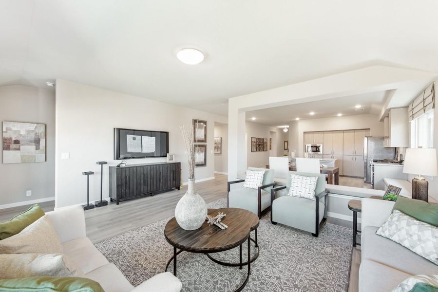 Open-concept living room with light gray sofas, round coffee table, wall-mounted TV, and kitchen visible in background Open-concept living room with light gray sofas, round coffee table, wall-mounted TV, and kitchen visible in background
