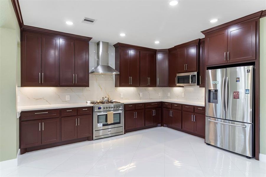 This kitchen features elegant dark wood cabinetry, stainless steel appliances, and a modern range hood. It has a spacious layout with sleek white countertops and tile flooring, providing a clean and contemporary feel.