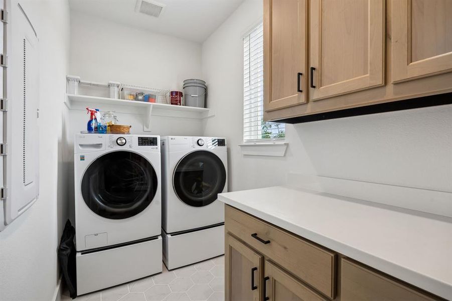 Laundry room featuring light wood-finish cabinetry with black hardware, white countertops, open shelving, and hexagonal floor tiling Laundry room featuring light wood-finish cabinetry with black hardware, white countertops, open shelving, and hexagonal floor tiling