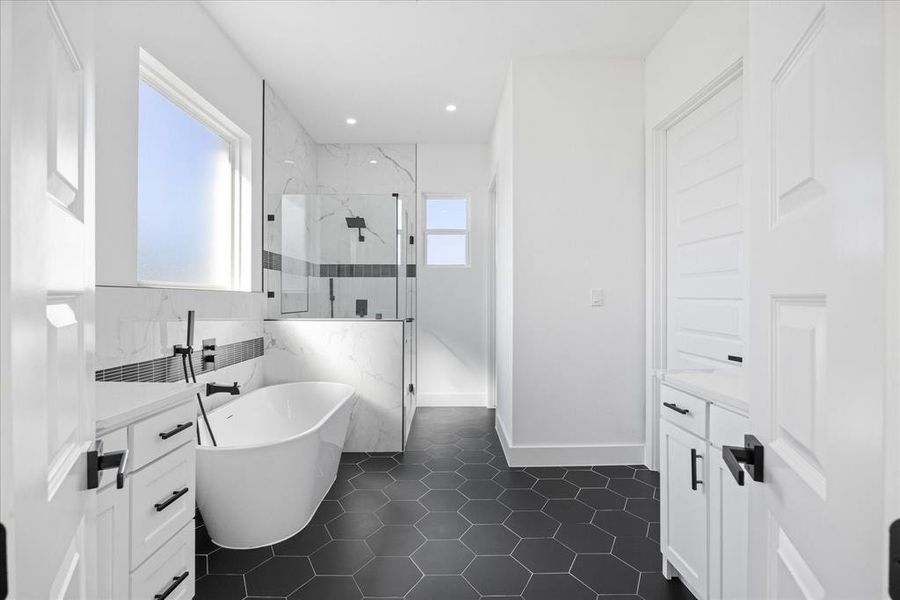 Modern bathroom featuring a freestanding tub with matte black fixtures, a spacious walk-in shower with clear glass enclosure, white cabinetry with matte black hardware, hexagon tile flooring, and a large window providing ample natural light
