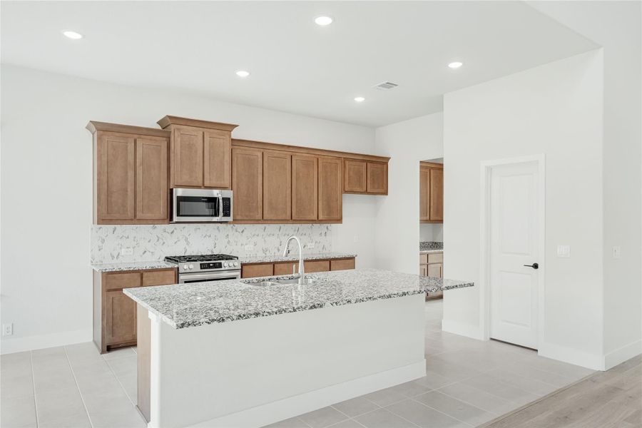 Kitchen with brown wood cabinets, granite island with sink, and stainless steel appliances on tile floor