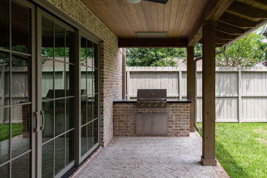 Covered back patio with wood ceiling, ceiling fan, brick exterior wall, and a built-in outdoor kitchen featuring a stainless steel grill, stone countertop, and stainless storage cabinet. Large multi-panel sliding glass doors open directly to the interior. Private fenced backyard with mature trees. Covered back patio with wood ceiling, ceiling fan, brick exterior wall, and a built-in outdoor kitchen featuring a stainless steel grill, stone countertop, and stainless storage cabinet. Large multi-panel sliding glass doors open directly to the interior. Private fenced backyard with mature trees.