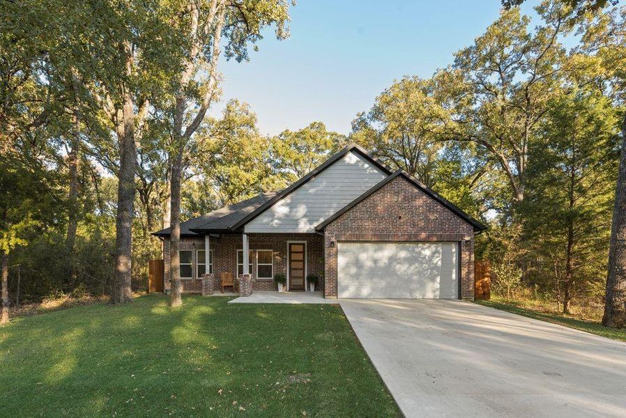 View of front facade featuring brick siding, a garage, driveway, a front yard, and view of wooded area