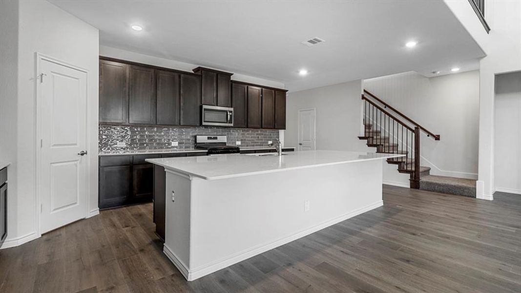 Spacious kitchen featuring an expansive island with a light-toned countertop, dark wood-finish cabinetry, a subway tile backsplash, and stainless steel appliances