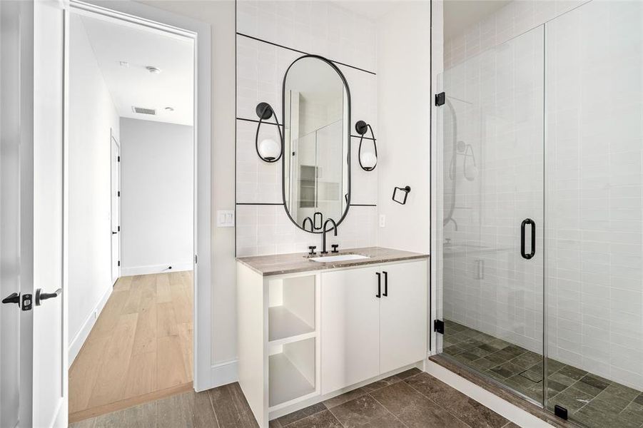 This ensuite bath exudes sophistication, featuring chic light fixtures, a bespoke mirror, luxurious stone counters and flooring, and a glass-enclosed tile walk-in shower, creating an elegant and serene retreat.