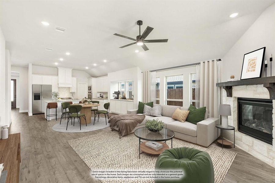 Open-concept living room with stone fireplace, ceiling fan, and kitchen visible in background on wood floors