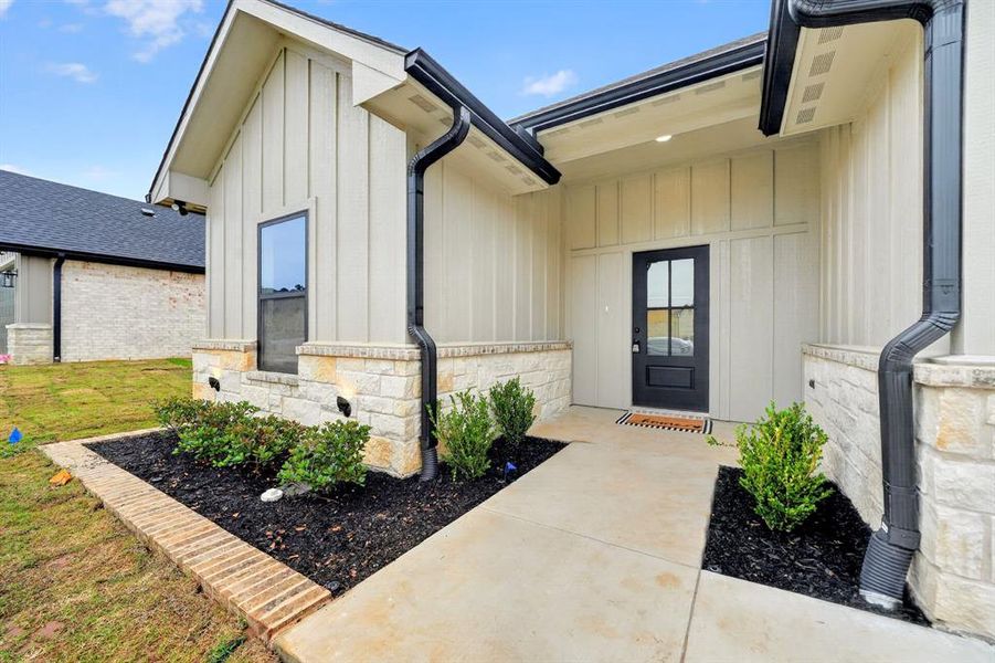 Contemporary exterior featuring board and batten siding, stone accents, a dark-framed glass-paneled entry door, black gutters and downspouts, and integrated landscaping