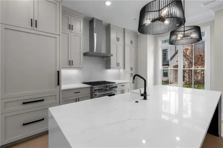 White stone countertop island with an undermount sink and matte black faucet, custom light gray cabinetry, stainless steel range with matching hood, and contemporary pendant lighting