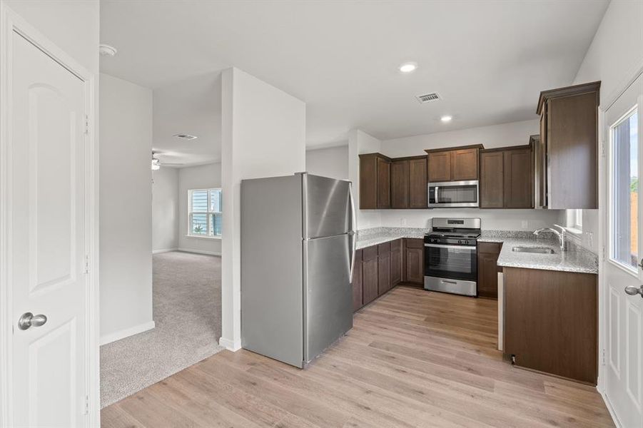 Kitchen with appliances with stainless steel finishes, dark brown cabinets, light stone countertops, light wood-style flooring, and a ceiling fan
