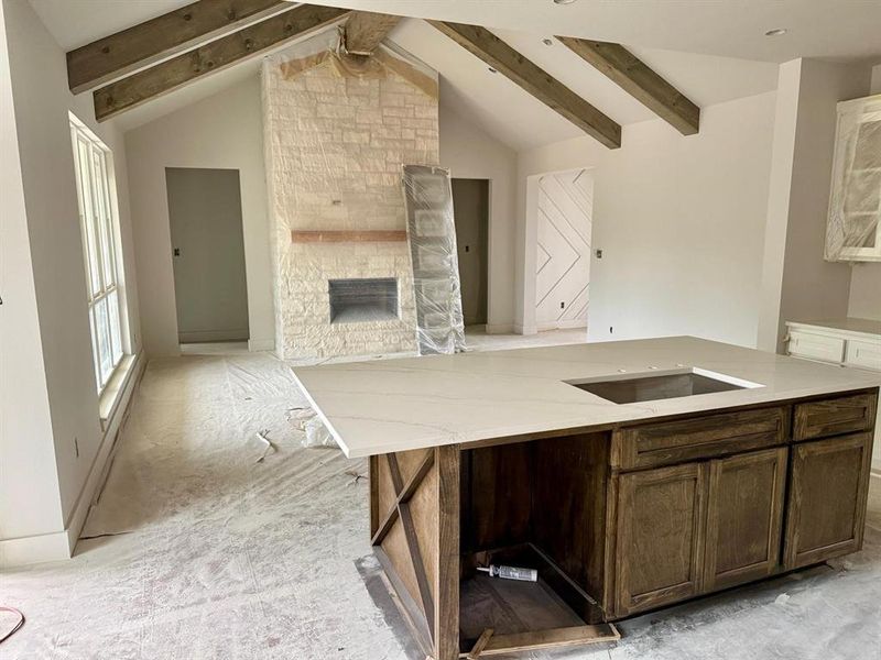Kitchen featuring an island with sink, dark brown cabinetry, a stone fireplace, light stone counters, and open floor plan Kitchen featuring an island with sink, dark brown cabinetry, a stone fireplace, light stone counters, and open floor plan