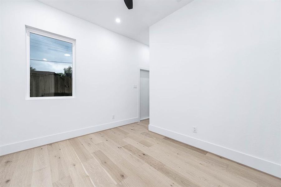 This is a clean, modern room with bright white walls, light wood flooring, and a single, central window providing natural light. It features a high ceiling with a ceiling fan, and ready for personalization.