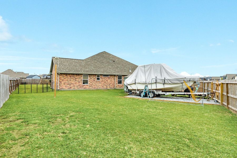 Spacious backyard with a well-maintained lawn, featuring a fenced perimeter. There's a covered boat on a trailer on a gravel pad, suggesting ample storage space. The house has a brick exterior and a sloped roof.