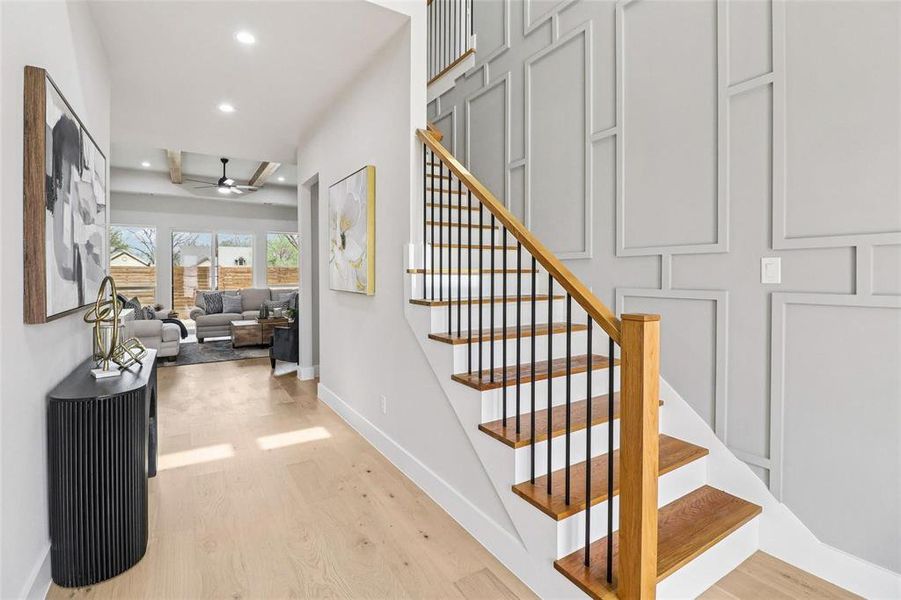 The inviting entryway features light-toned wood flooring, a staircase with wooden treads and a black metal railing, and decorative wall paneling