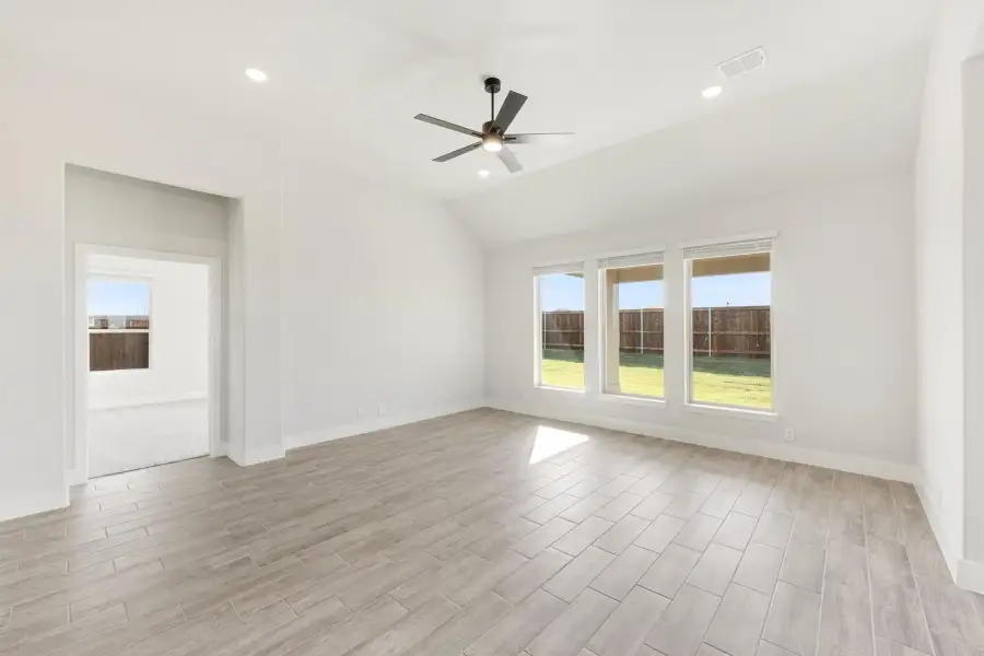 Empty living room with vaulted ceiling, ceiling fan, wood-look tile floors, and large windows overlooking backyard
