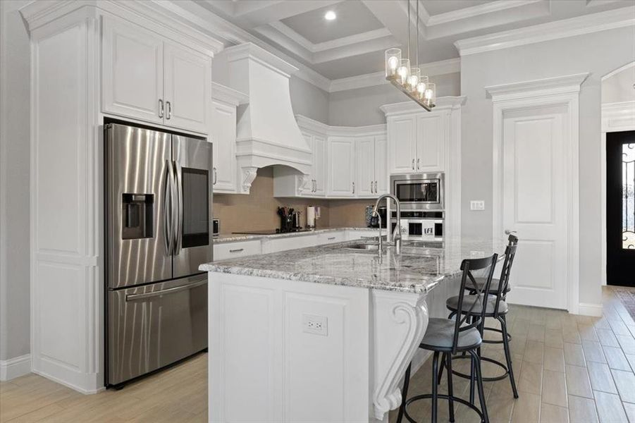 Kitchen featuring light stone counters, a kitchen island with sink, premium range hood, white cabinets, and appliances with stainless steel finishes