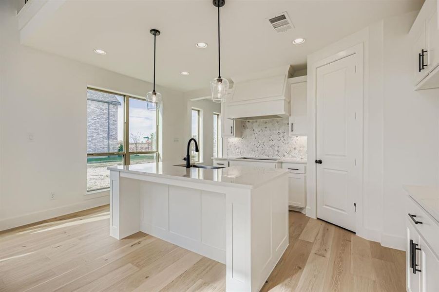 Kitchen featuring tasteful backsplash, a kitchen island with sink, white cabinets, light stone counters, and decorative light fixtures Kitchen featuring tasteful backsplash, a kitchen island with sink, white cabinets, light stone counters, and decorative light fixtures