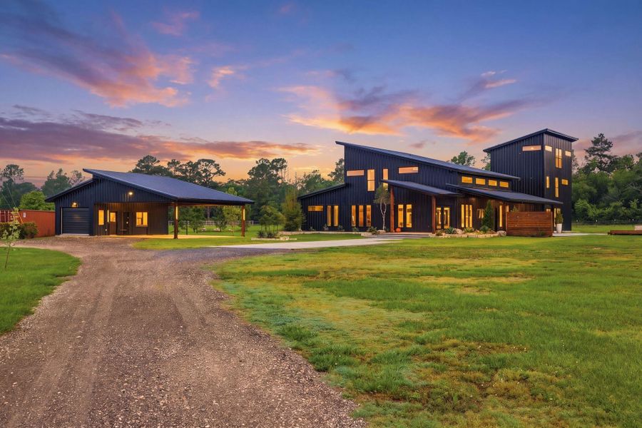 Modern, spacious home with a sleek, dark exterior and large windows, set on a sizable, green lot. A long driveway leads to both the house and a detached carport with versitile shop space- laundry room and full bathroom.