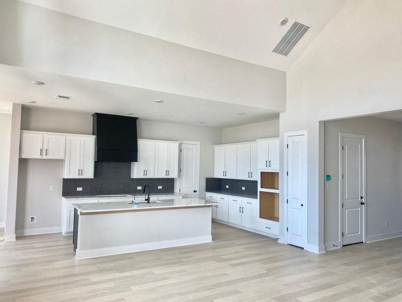 Kitchen featuring white cabinets, backsplash, a kitchen island with sink, light wood-style floors, and a towering ceiling Kitchen featuring white cabinets, backsplash, a kitchen island with sink, light wood-style floors, and a towering ceiling