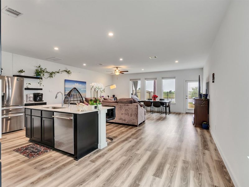 This open-concept living area features modern finishes, including wood-style flooring and recessed lighting. The sleek kitchen has stainless steel appliances and a large island. The space flows into a cozy seating area and a dining space with natural light from large windows, offering a bright and inviting atmosphere.