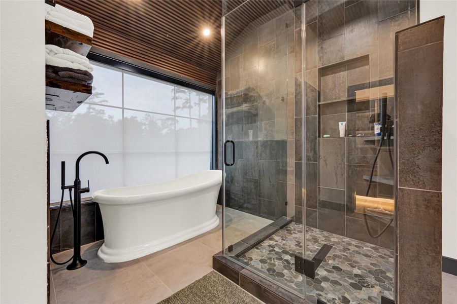 Freestanding bathtub next to a spacious glass-enclosed shower with stylish dark tiles and a pebble stone floor. A wood-paneled ceiling and frosted window add warmth and privacy to the space.
