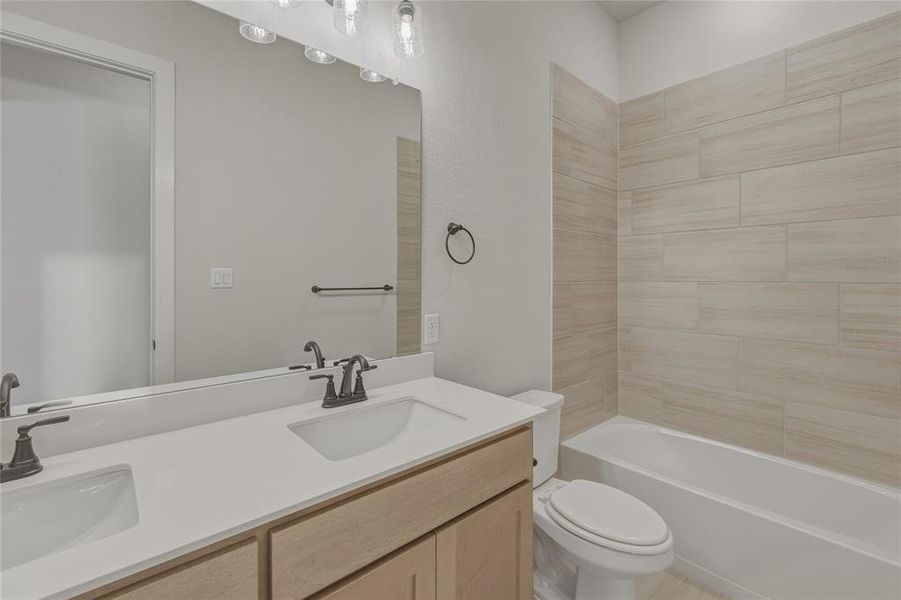Bathroom featuring a double vanity with light wood-finish cabinetry and white countertops, dual undermount sinks with bronze-finish fixtures, a large frameless mirror, and a tiled shower-tub combination Bathroom featuring a double vanity with light wood-finish cabinetry and white countertops, dual undermount sinks with bronze-finish fixtures, a large frameless mirror, and a tiled shower-tub combination