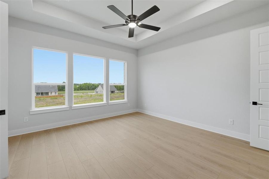 This room features a tray ceiling with a ceiling fan, light-colored flooring, and large windows that provide views of undeveloped land and distant buildings