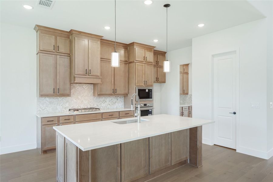 Kitchen with light wood cabinets, large island with white countertop, herringbone backsplash, and pendant lights
