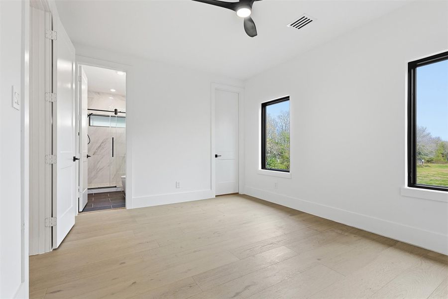 The bedroom features light-toned flooring, a matte black ceiling fan, and dual black-framed windows that provide ample natural light.