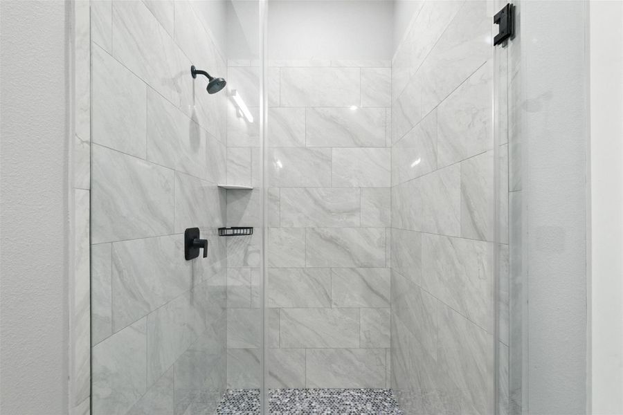 This photo showcases a modern shower with elegant marble-style tiles and a sleek glass door. The fixtures are a stylish matte black, and the flooring features a contrasting pebble-like texture, providing a contemporary and inviting feel.