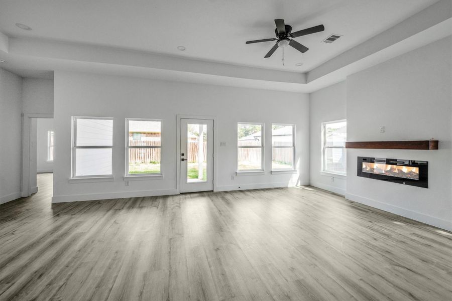 This spacious living room features a modern electric fireplace, abundant natural light from multiple windows, and sleek wood flooring. The ceiling fan adds comfort, and the room opens to a private backyard through a glass door.
