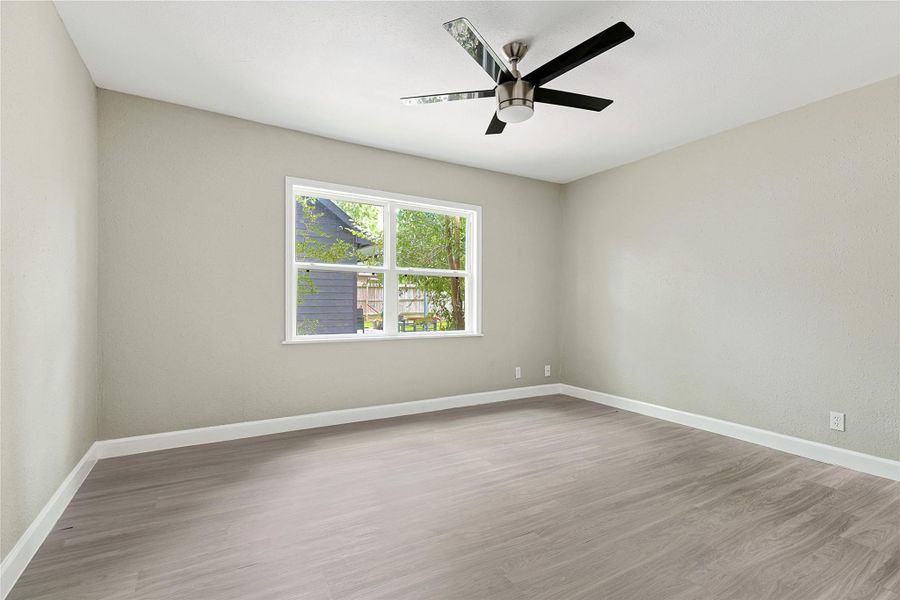 This room features light gray walls, wood flooring, a ceiling fan, and a large window that offers natural light and a view of greenery outside. Perfect as a bedroom or office space.