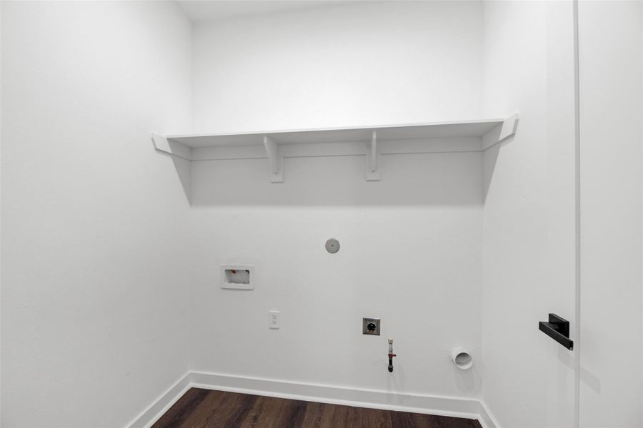 Laundry room with white walls and a dark wood floor. It features utility hookups and has a single wall-mounted shelf for storage.