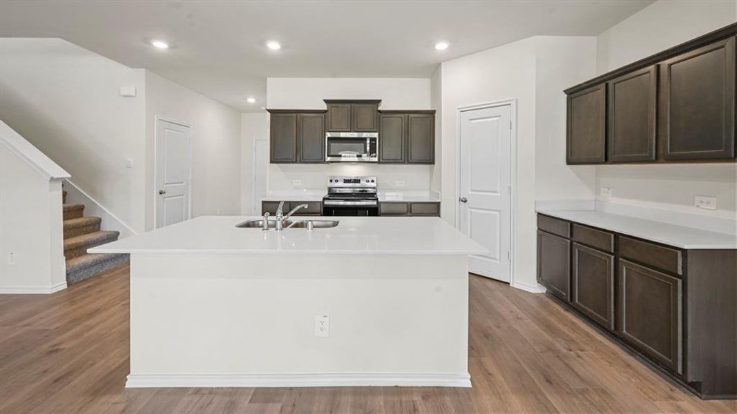 Modern kitchen featuring a large white island with an undermount sink, dark wood-finish cabinetry, stainless steel appliances, white countertops, and wood-finish flooring