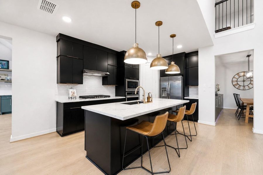 This modern kitchen features sleek cabinetry with hardware, stainless steel appliances, and a spacious quartz island with bar seating. Pendant lighting adds warmth, while an open layout connects to a dining area, enhancing the space's airy feel.