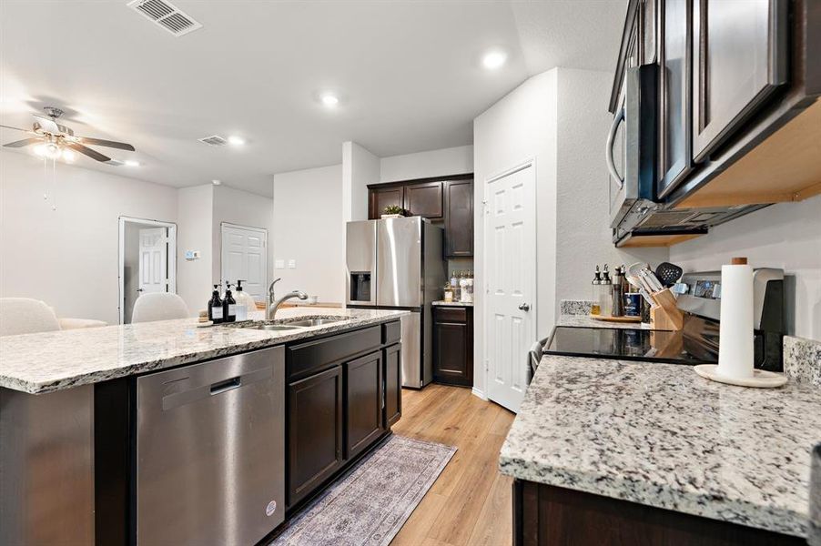 Kitchen featuring light stone counters, appliances with stainless steel finishes, a center island with sink, dark brown cabinetry, and recessed lighting Kitchen featuring light stone counters, appliances with stainless steel finishes, a center island with sink, dark brown cabinetry, and recessed lighting