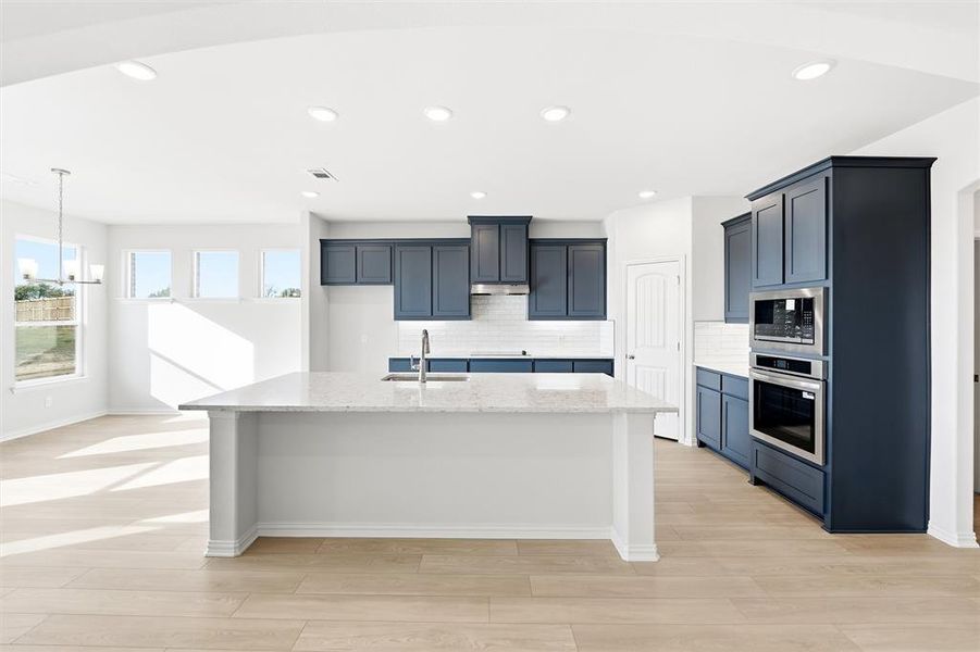 Kitchen with appliances with stainless steel finishes, light stone countertops, an island with sink, decorative backsplash, and light wood finished floors