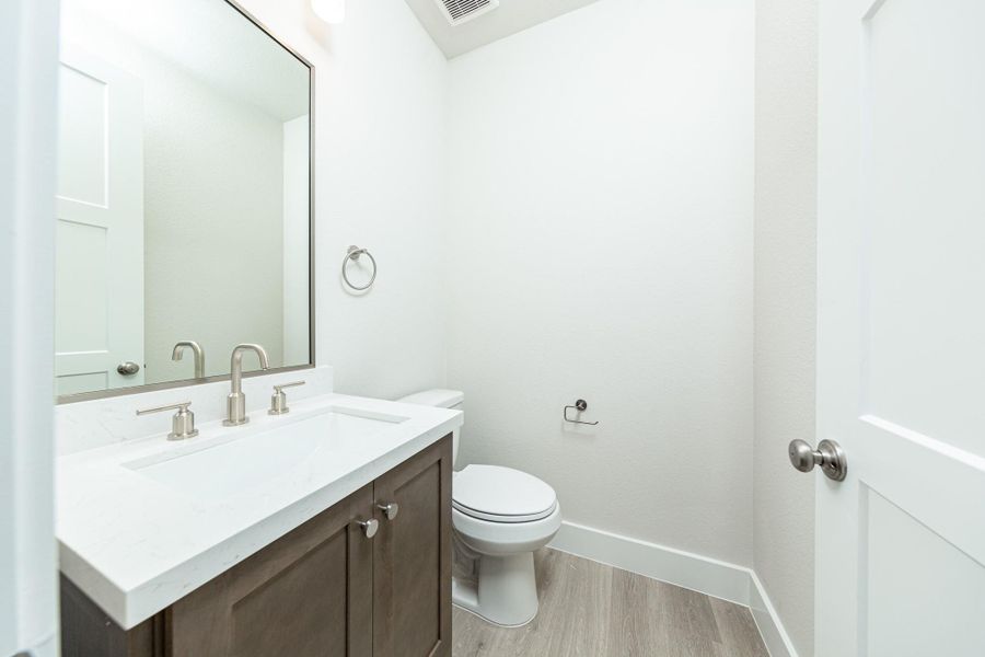 The first-floor powder room offers convenience and a modern aesthetic. It features a stained cabinet, quartz countertop, brushed nickel sleek faucet and a matching wall-mounted towel ring, laminate flooring, and a framed mirror.