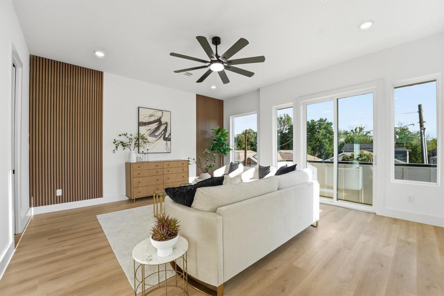 Bright, contemporary living room featuring wood-look flooring, recessed lighting, a modern ceiling fan, accent wood paneling, and expansive sliding glass doors that fill the space with natural light and provide access to a private balcony.